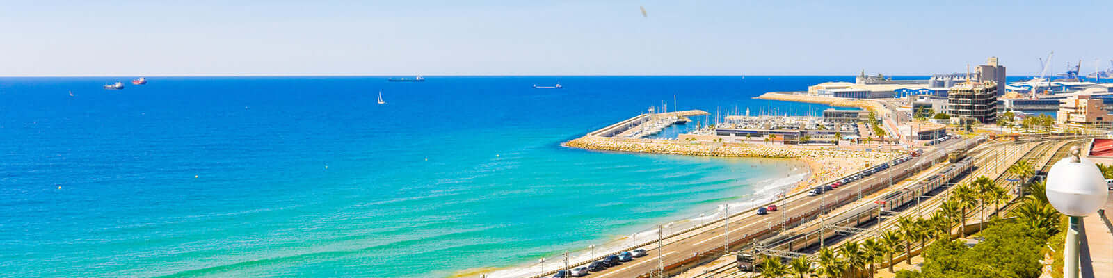 A scenic coastal view featuring a vibrant blue sea, with a marina and sandy beach stretching alongside a road lined with palm trees. Several ships are visible on the horizon under a clear blue sky.