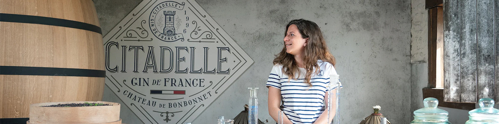 A woman in a striped shirt stands at a counter with bottles and a large cask behind her. The wall features the text Citadelle Gin de France with a logo.