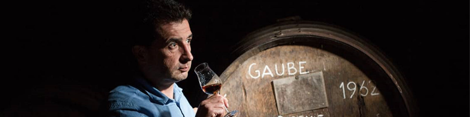 A person in a blue shirt holds a wine glass thoughtfully in front of a large wooden barrel labeled Gaube 1982 in dim lighting.