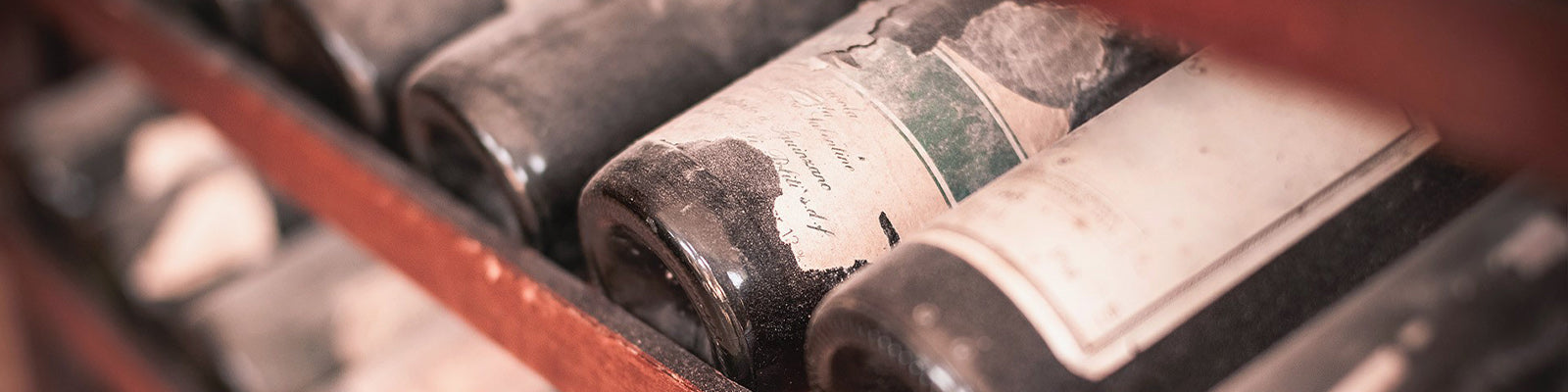 Close-up of dusty wine bottles lying on their sides in a wooden rack. The bottles have old, faded labels, suggesting they have been stored for a long time.