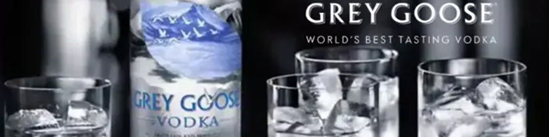 Bottiglia di vodka Grey Goose accanto a tre bicchieri pieni di ghiaccio e vodka, su uno sfondo scuro. Il testo recita Grey Goose Worlds Best Tasting Vodka.