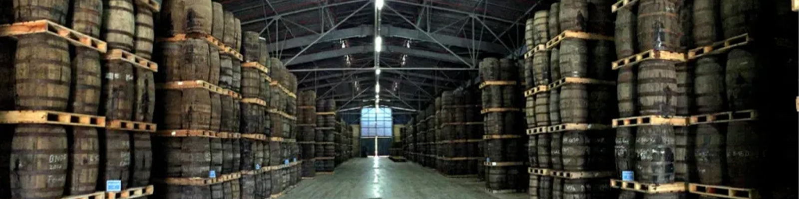 A warehouse filled with rows of stacked wooden barrels on pallets, under a high, arched metal ceiling with overhead lights. The scene is symmetrical and spacious, providing an industrial feel.