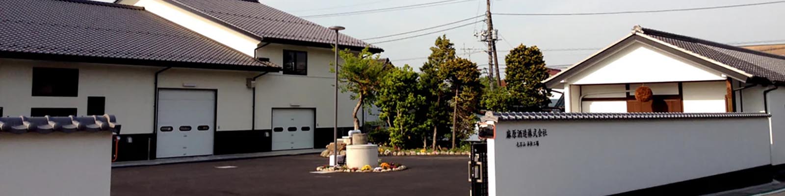 A modern Japanese building with traditional architectural elements, featuring a tiled roof and white walls. A well-manicured garden and trees are visible, along with a stone pathway and sign with Japanese text.