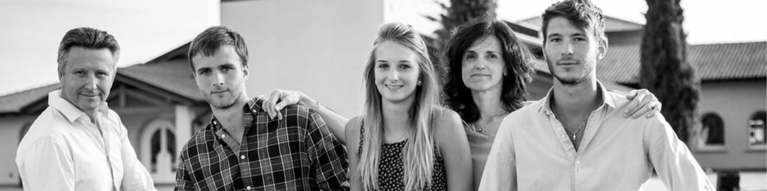 A black and white photo of a group of five people standing close together outside a building. They are smiling and casually dressed, with two men on each side and a woman in the center.