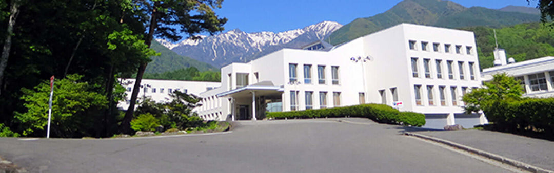 A white building with multiple windows sits at the end of a curved driveway. Its surrounded by lush greenery and tall mountains in the background under a clear blue sky.