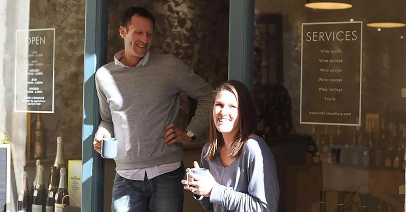 Ed and Jess standing outside a store with blue doors, holding mugs and smiling, with wine bottles displayed near the entrance