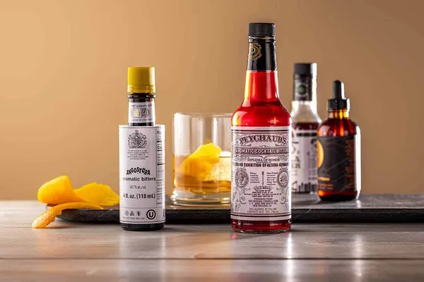 Bottles of cocktail bitters on a wooden table beside a glass with a slice of orange peel