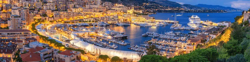 A marina at dusk with numerous yachts docked in the harbour and the city lit behind