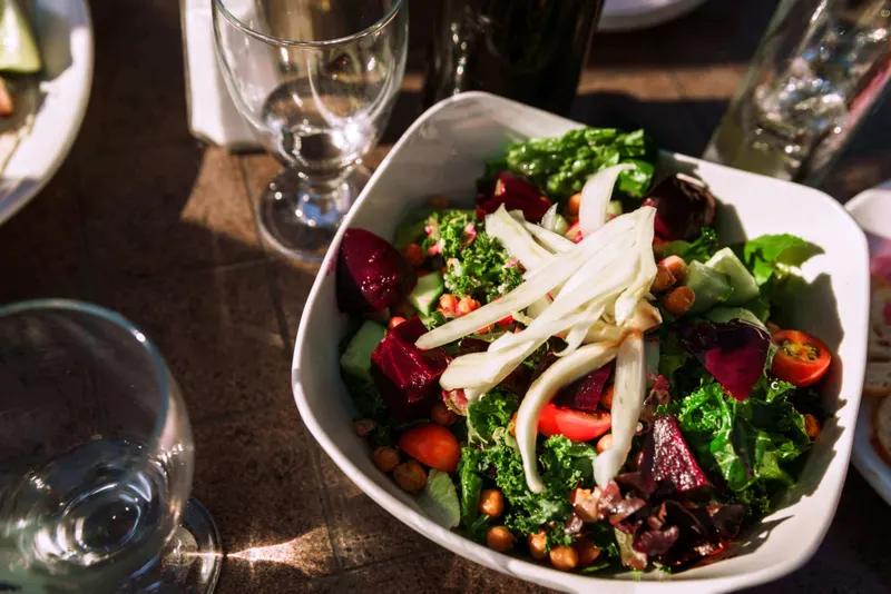 A sunlit wooden table set for lunch with a salad, wine glasses and bottles