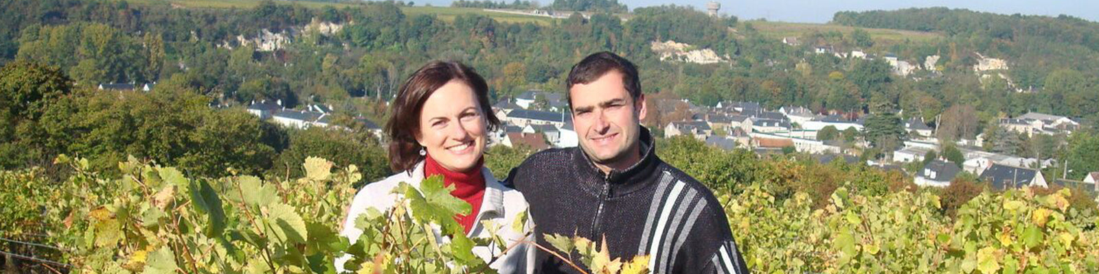 A man and a woman stand together in a vineyard, with a scenic view of a village and hills in the background. They are smiling and dressed in sweaters on a sunny day.