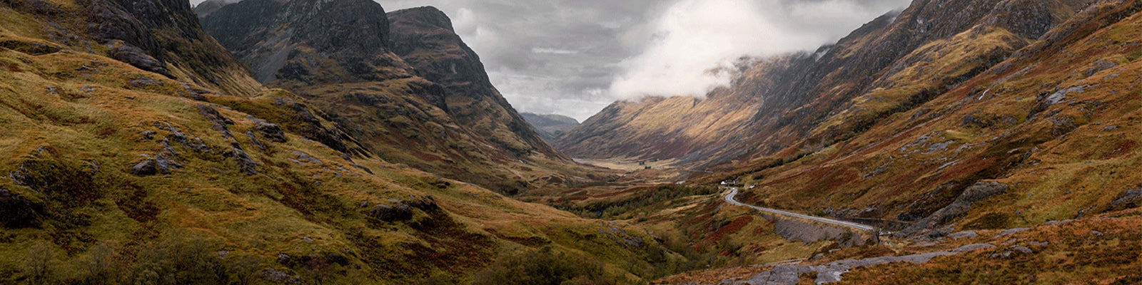 A panoramic view of a misty valley between rugged, green and brown mountains. A narrow road winds through the landscape, with cloudy skies overhead.