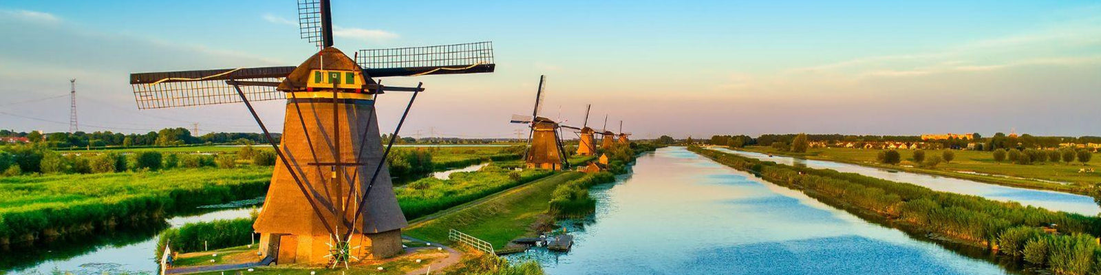 A scenic view of traditional windmills along a tranquil river in a lush, green landscape during sunset. The sky is a soft gradient of blue and orange, reflecting on the waters surface.