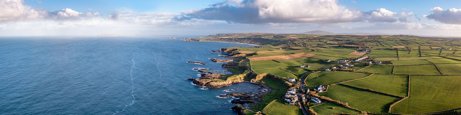 A panoramic view of a coastal landscape with vast green fields meeting the rocky shoreline. The ocean stretches into the horizon under a partly cloudy sky, with scattered houses dotting the countryside.