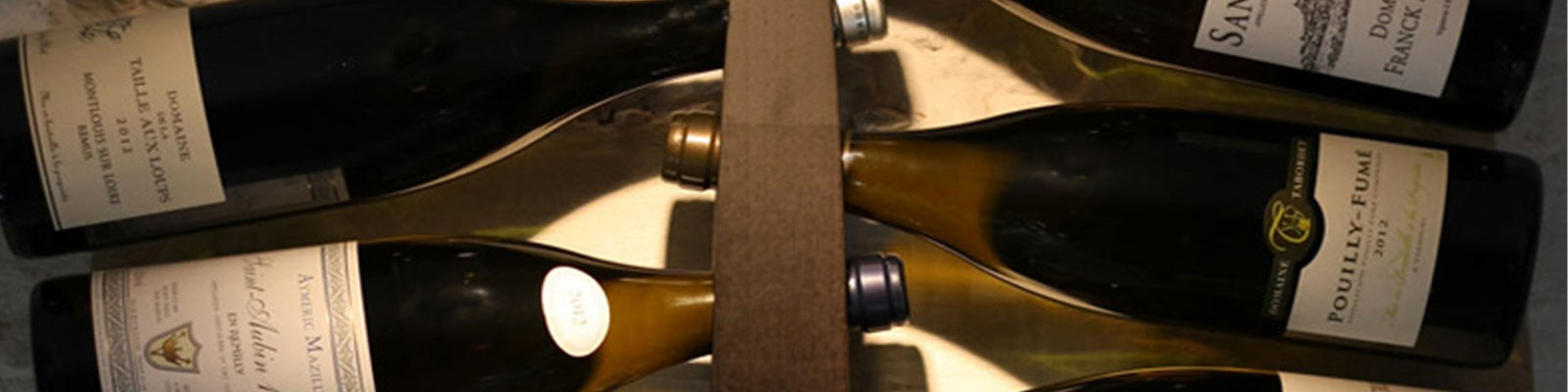 Close-up view of five wine bottles lying horizontally in a wooden rack. Labels on the bottles include Pouilly-Fumé and Sancerre, indicating a collection of white wines. The lighting casts a warm glow on the labels.