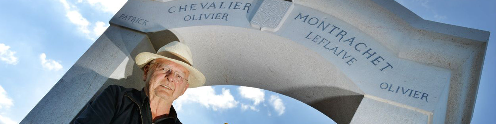 A man in a hat stands under a stone arch with engraved names such as Chevalier Montrachet and Leflaive. The sky is bright blue with some clouds in the background.