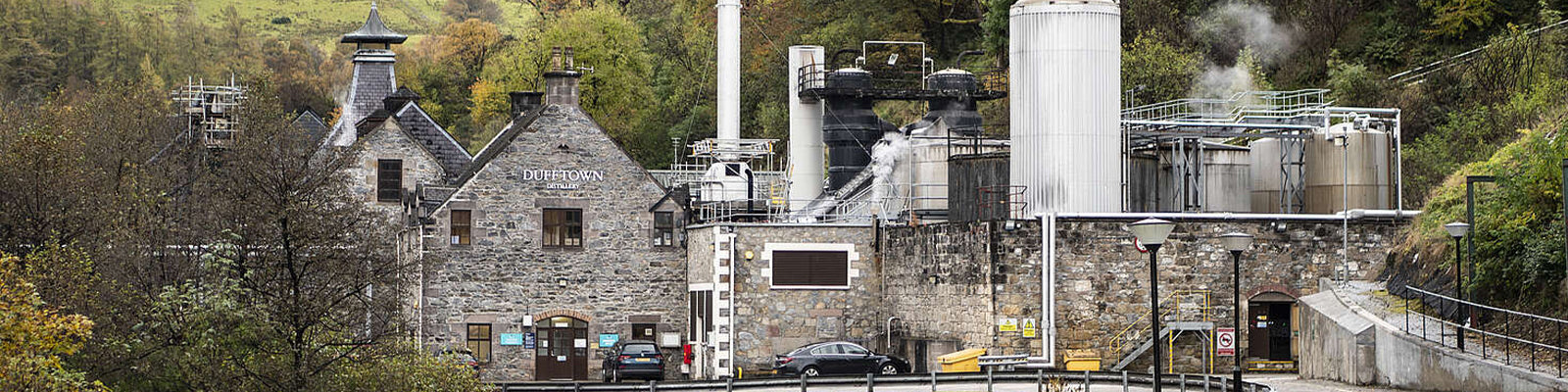 A distillery with historic stone buildings and modern industrial structures, surrounded by trees and hills. Steam rises from large metal tanks, and a few cars are parked outside the main building.