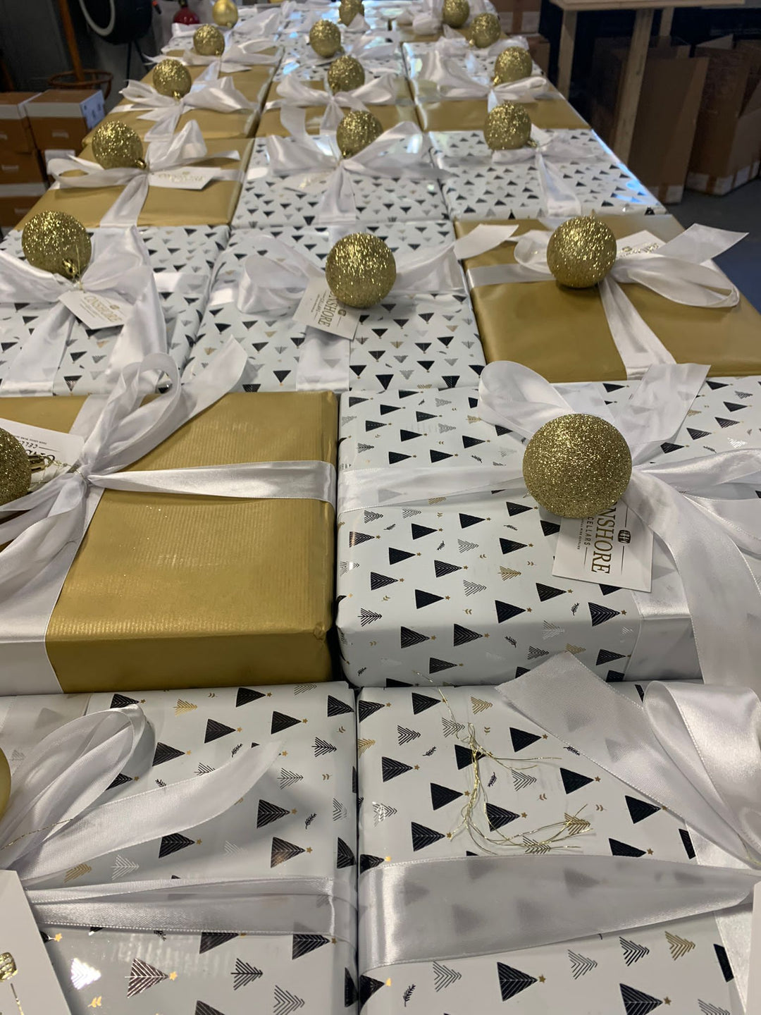 Rows of neatly wrapped gifts with white and black triangle-patterned paper, topped with white ribbons and golden glittery ornaments. Some gifts are wrapped in gold paper.